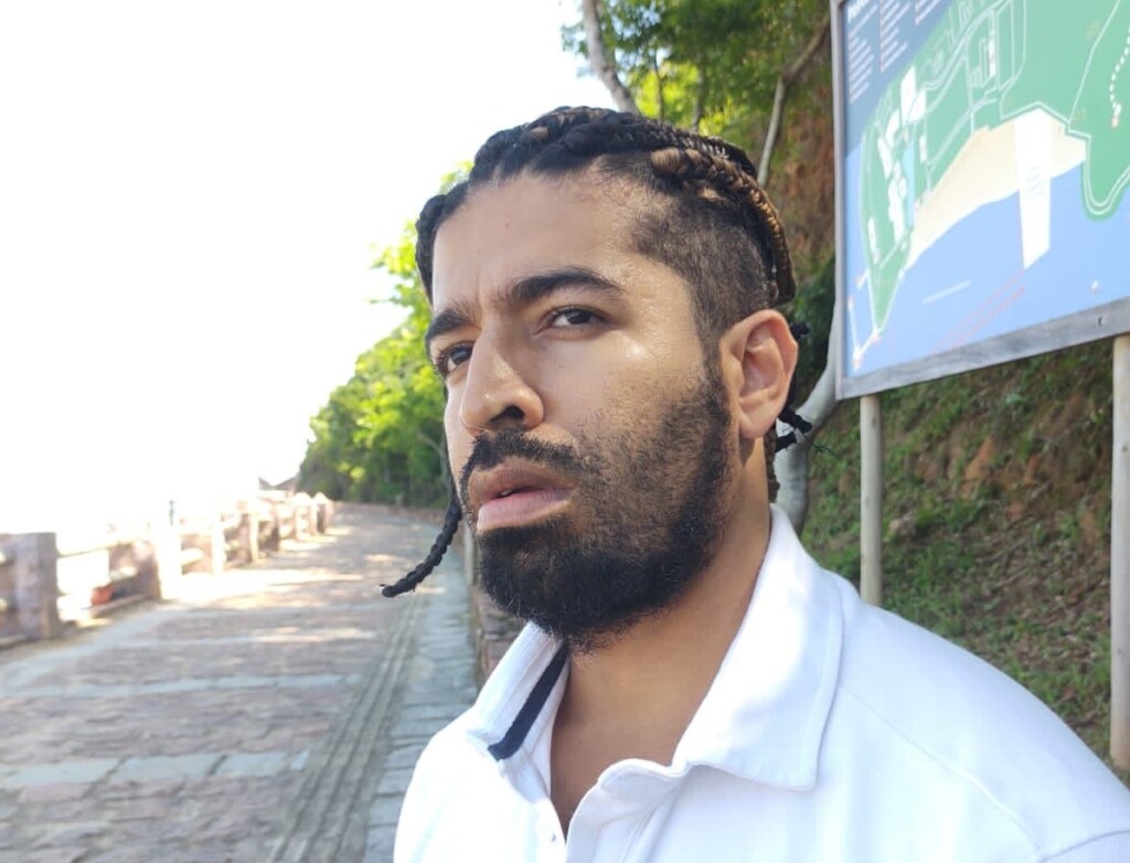Autor do livro Racismo Linguístico. Foto lateral com rosto do escritor. Ele é jovem, negro, tem cabelo trançado e barba. Veste camisa gola polo branca. 